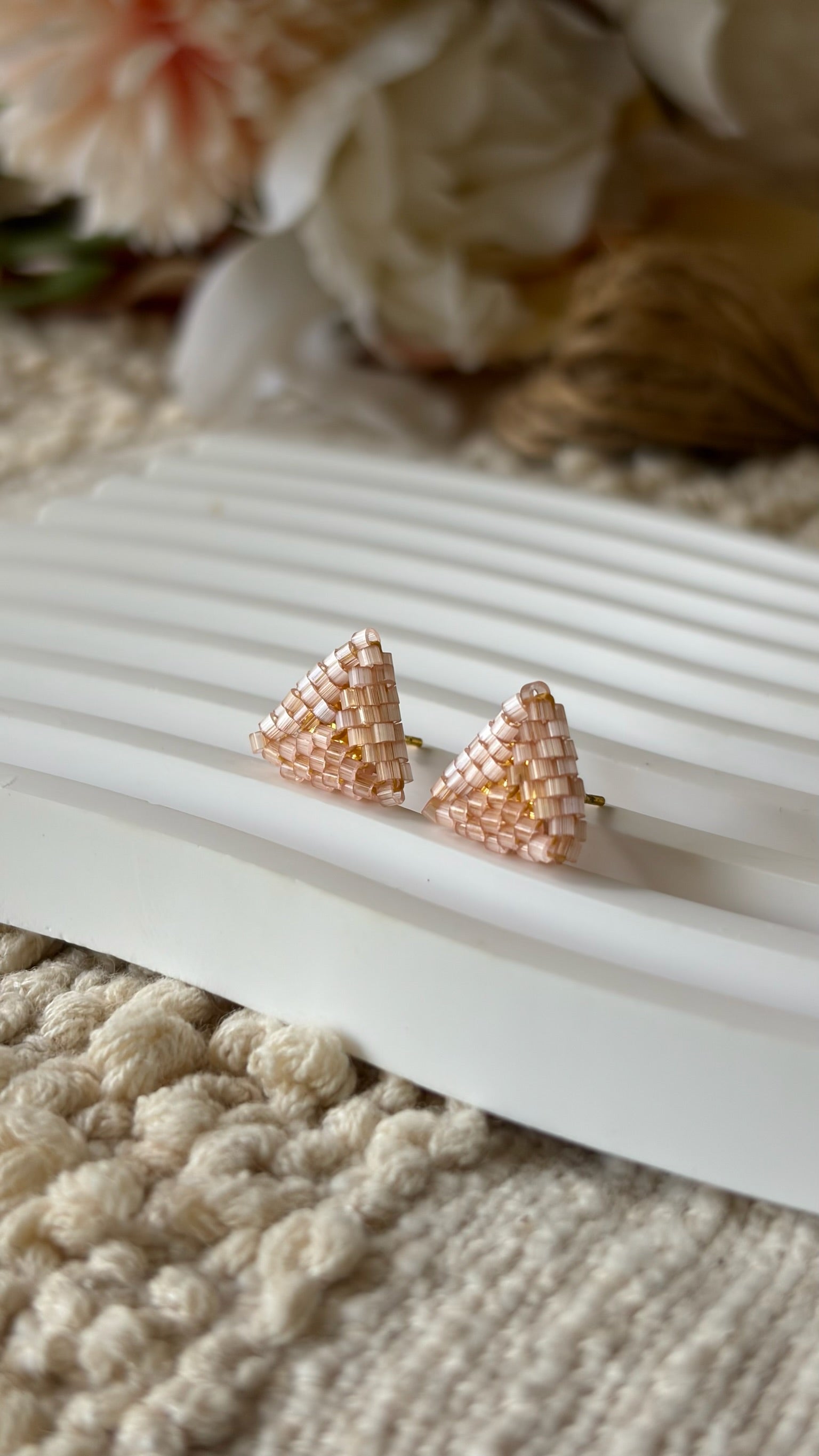 Rose gold triangle-shaped earrings on a textured white surface with beige flowers in the background.